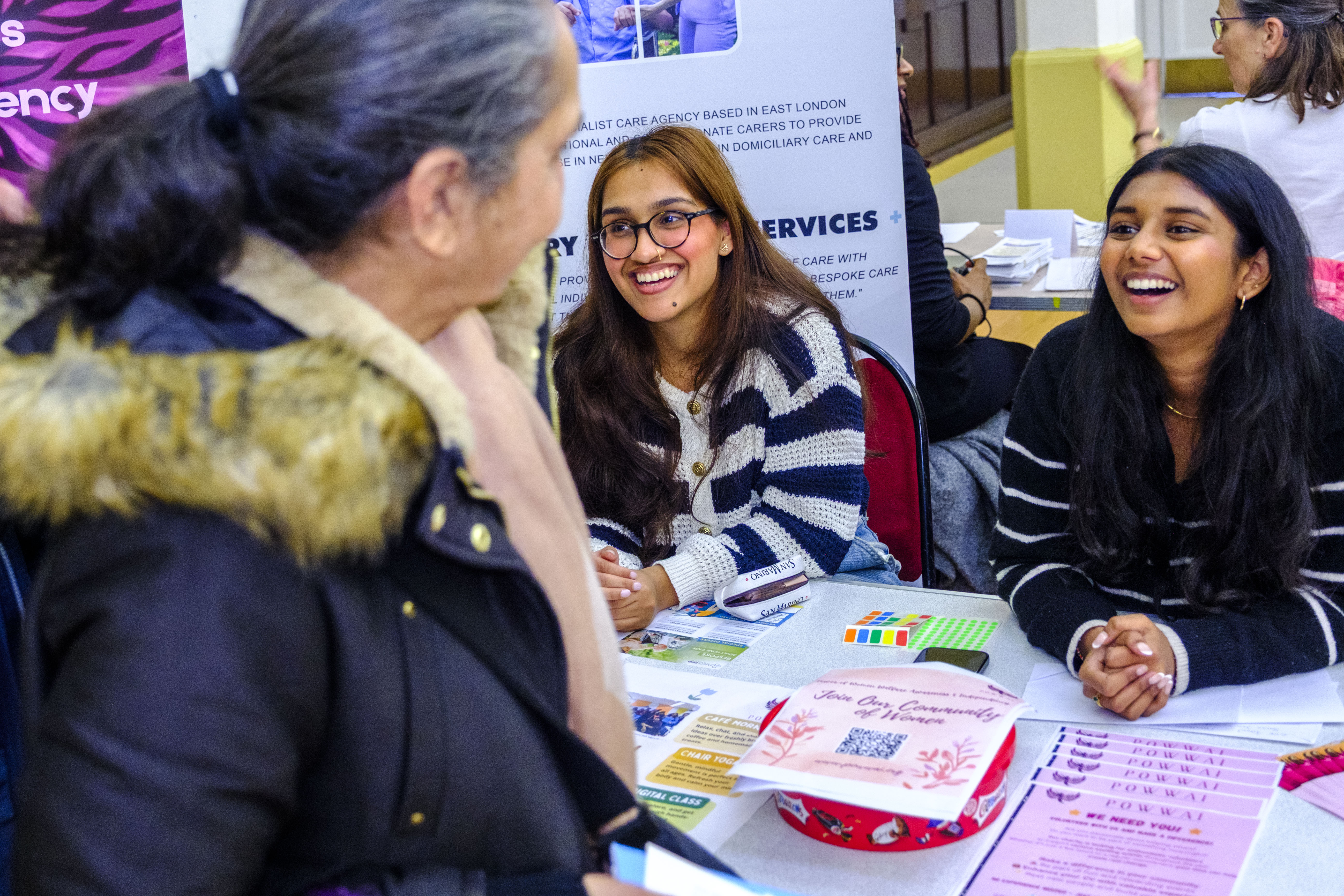 A photo of two young women sat at an information stall. They are smiling naturally at a person who is out of frame. Another woman walks past the stall. 
