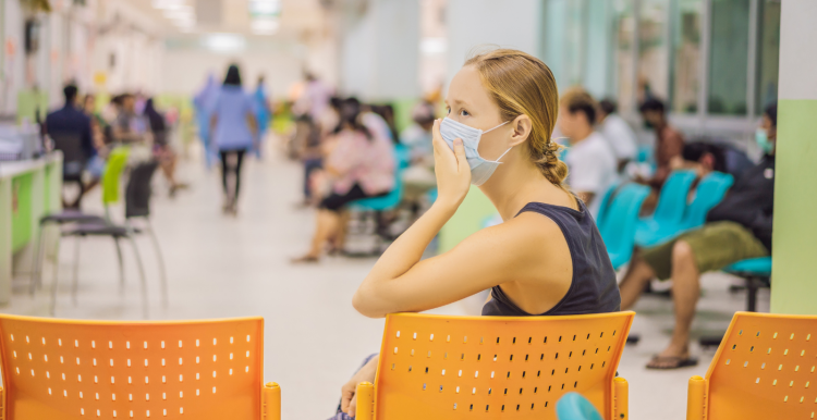 A photo of a woman in a hospital waiting room, she holds her hand to the mask covering her face and looks at something off screen nervously.