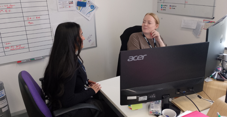 A photo of two women at a desk speaking to each other