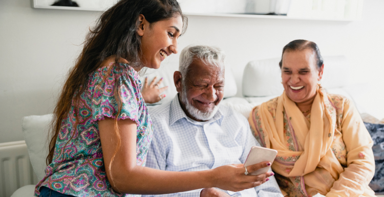 A photo of a young woman sat on a sofa next to an older couple, the young woman shows them something on her phone as they all smile.