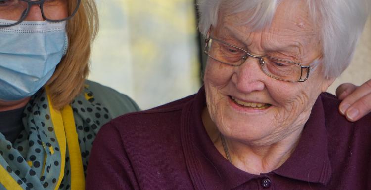 Smiling female carer in blue hospital mask alongside elderly female service user