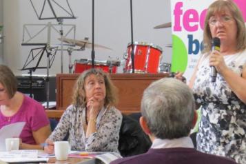 A photo of a woman in a conference space addressing members of a meeting.