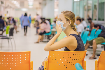 A photo of a woman in a hospital waiting room, she holds her hand to the mask covering her face and looks at something off screen nervously.