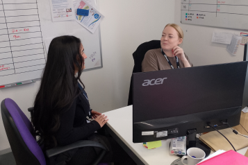 A photo of two women at a desk speaking to each other