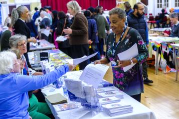 A photo of a busy hall with information stalls. A woman sat behind a desk offers a leaflet to a woman who is standing. 