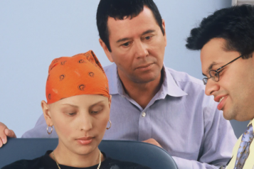 A woman with no hair wearing a head scarf, with her husband standing behind her