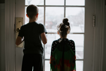 A boy and girl inside a house lookinhg out through the glass door onto the street