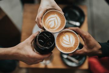 3 people in a cafe holding their hot drinks together - 'toasting' each other