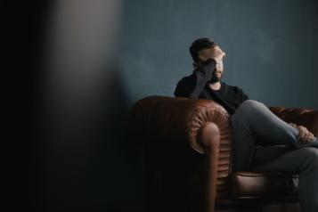 Young man sat on sofa resting head on his hand, looking despondent  