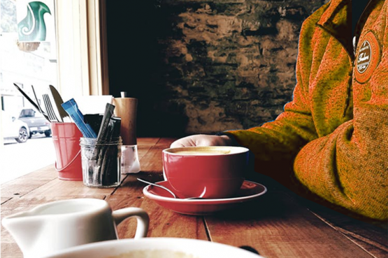 A person in an orange top sitting at a table in a cafe having a cup of coffee
