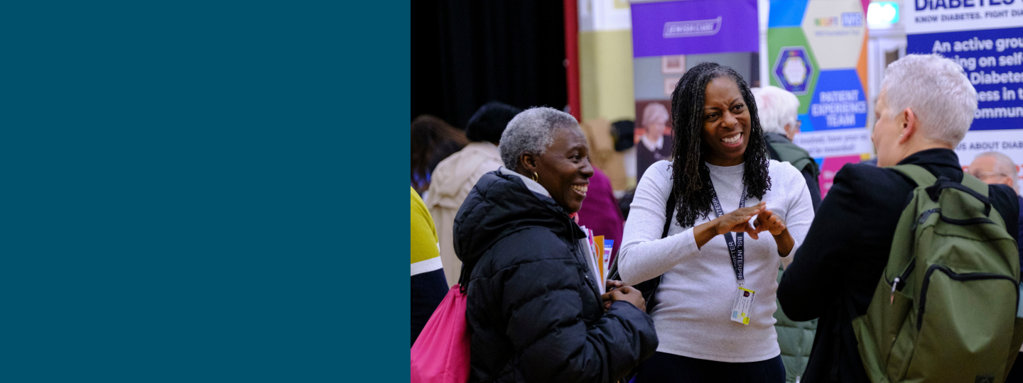 A photo of 3 people stood in a large public hall surrounded by community stalls. A woman in the middle communicates to the group with British Sign Language. 