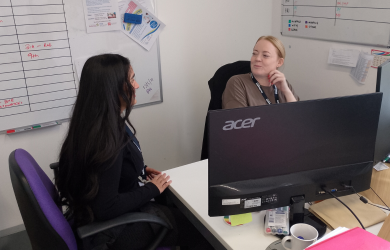 A photo of two women at a desk speaking to each other
