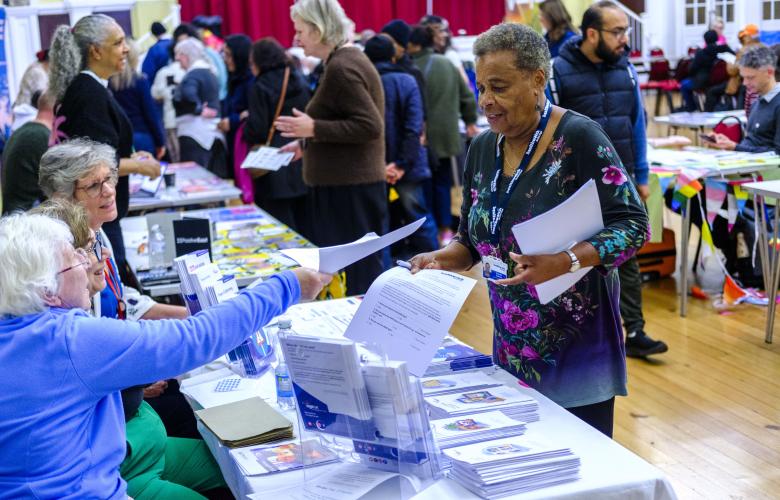 A photo of a busy hall with information stalls. A woman sat behind a desk offers a leaflet to a woman who is standing. 
